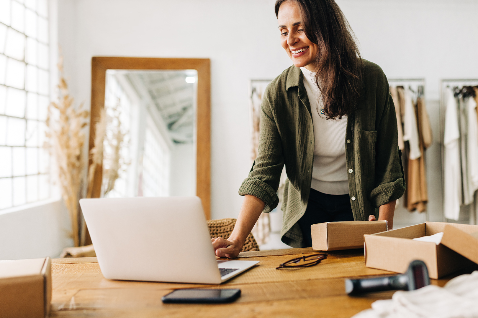 Eine lächelnde Frau arbeitet an einem Laptop, während sie Pakete in einem hellen Mode- oder Online-Shop vorbereitet.
