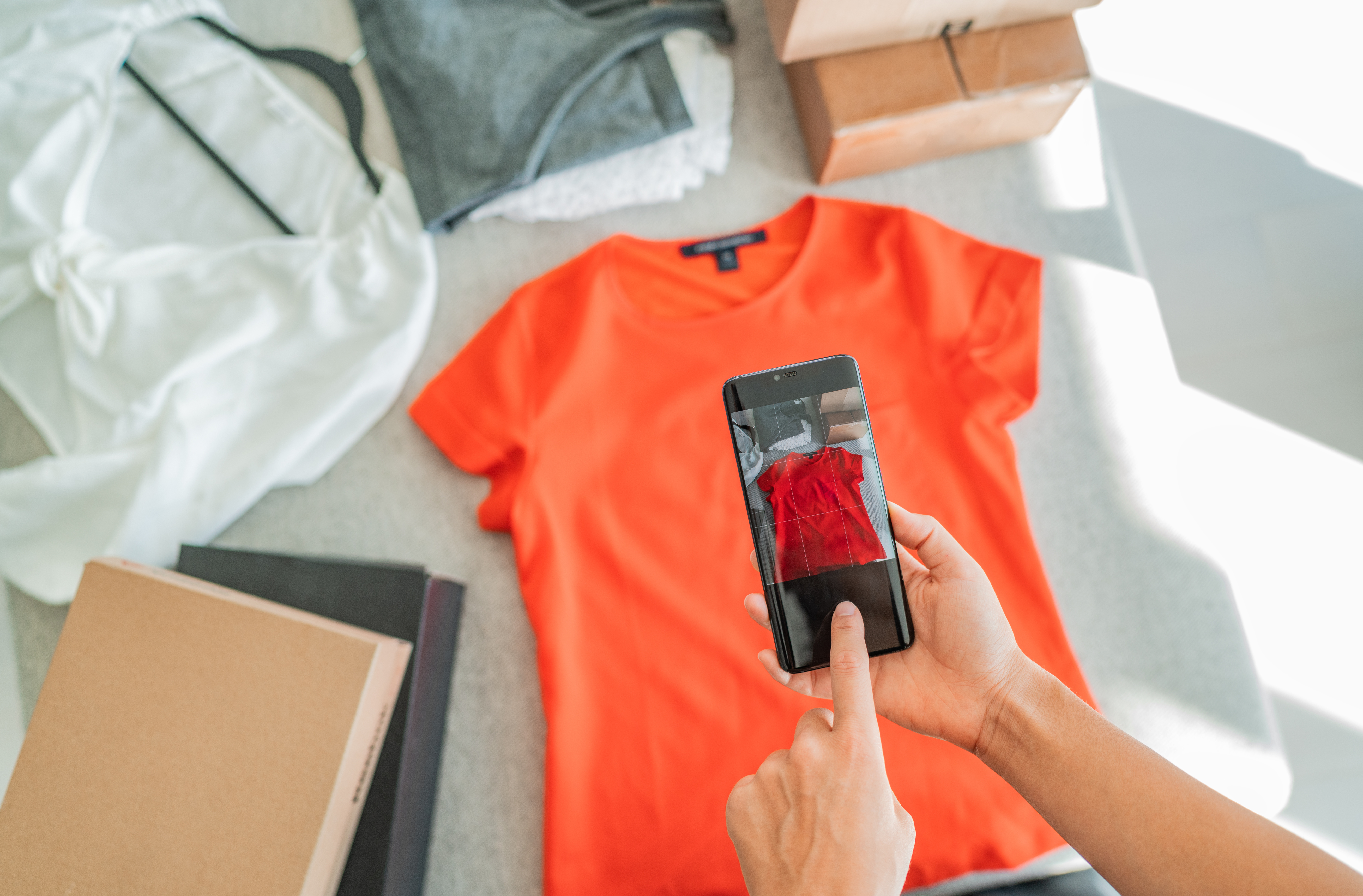 Eine Person fotografiert ein orangefarbenes T-Shirt mit dem Smartphone, um Produktbilder für einen Mode-Webshop zu erstellen.