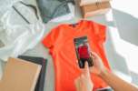 Eine Person fotografiert ein orangefarbenes T-Shirt mit dem Smartphone, um Produktbilder für einen Mode-Webshop zu erstellen.