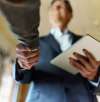 A businessman in a suit shaking hands with another person, holding a tablet, symbolizing a successful business agreement