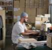 Man in a hat and glasses working on a laptop in a clothing webshop, surrounded by boxes and shoes